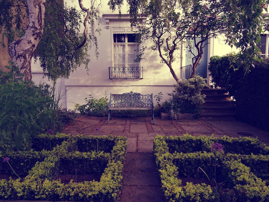 A small urban garden in Pimlico, featuring a paved patio area with a decorative wrought-iron garden bench centered against a white exterior wall of a building with a window and a small balcony. The ga